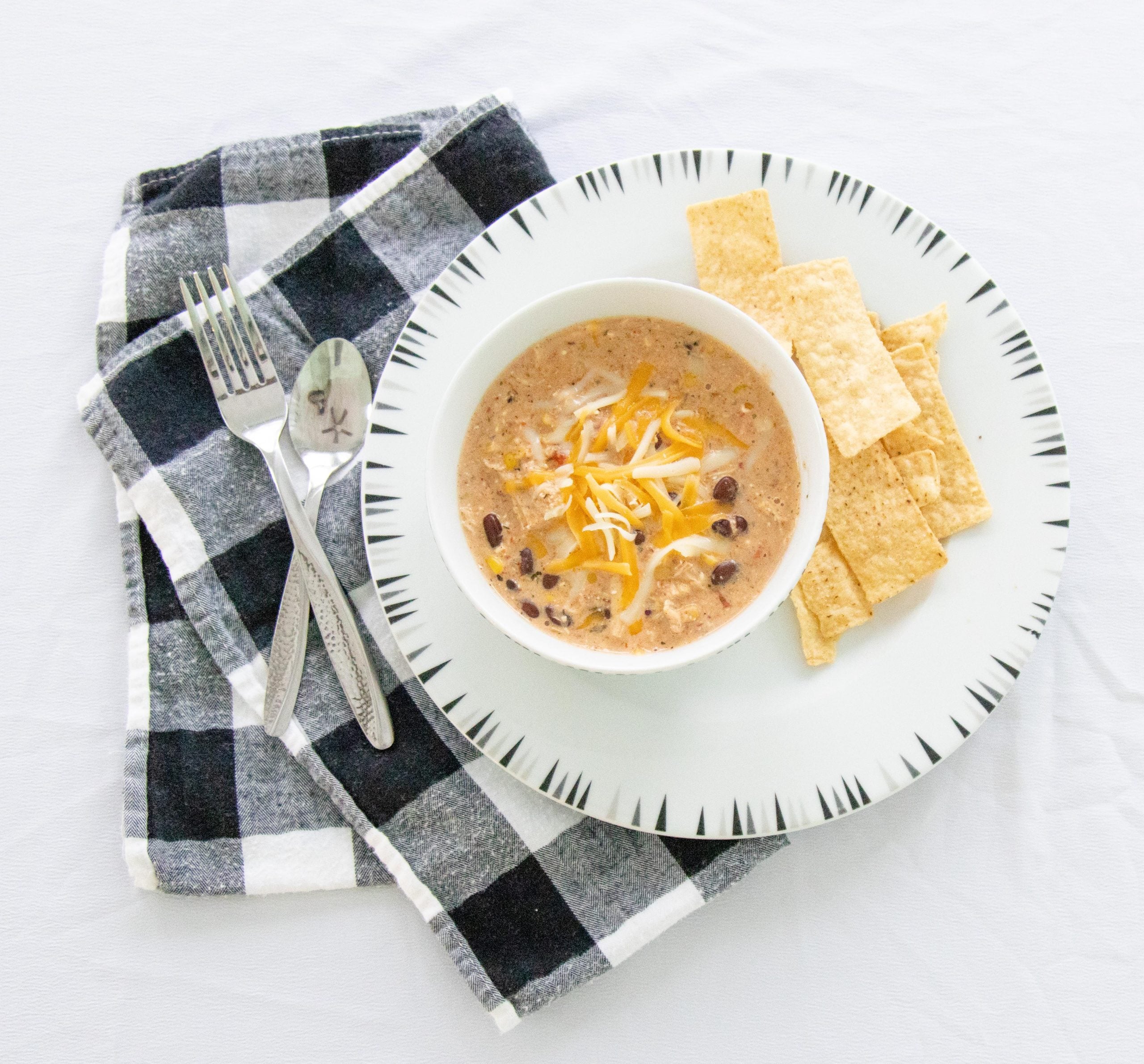 black and white checkered napkin with a white plate containing tortilla chips and a bowl of chicken chili soup