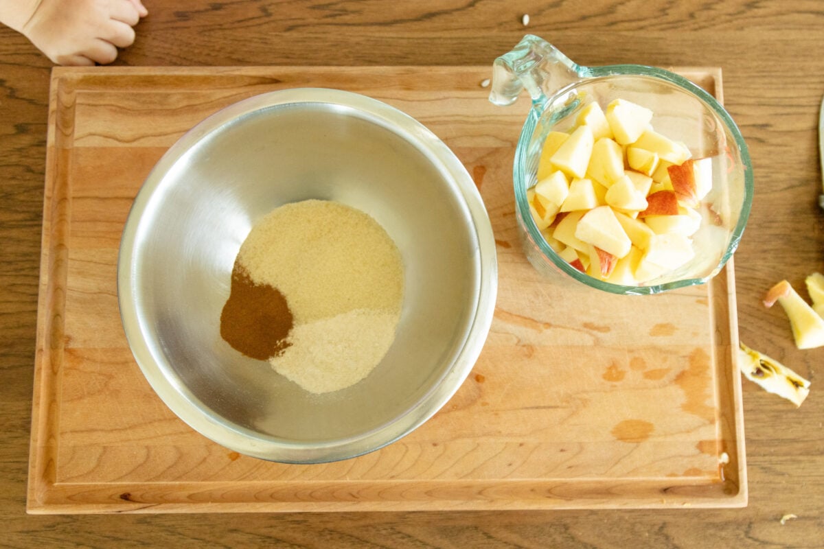 a container of apples and a mixing bowl with sugar and cinnamon in it setting on the cutting board